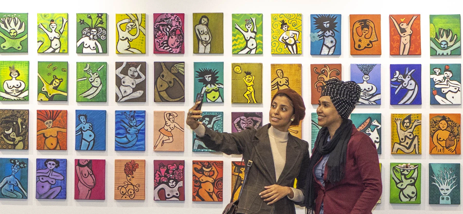 Zwei Besucherinnen machen ein Selfie vor einer Wand mit bunten Bildern aus der Werkreihe »Historias de Evas«.